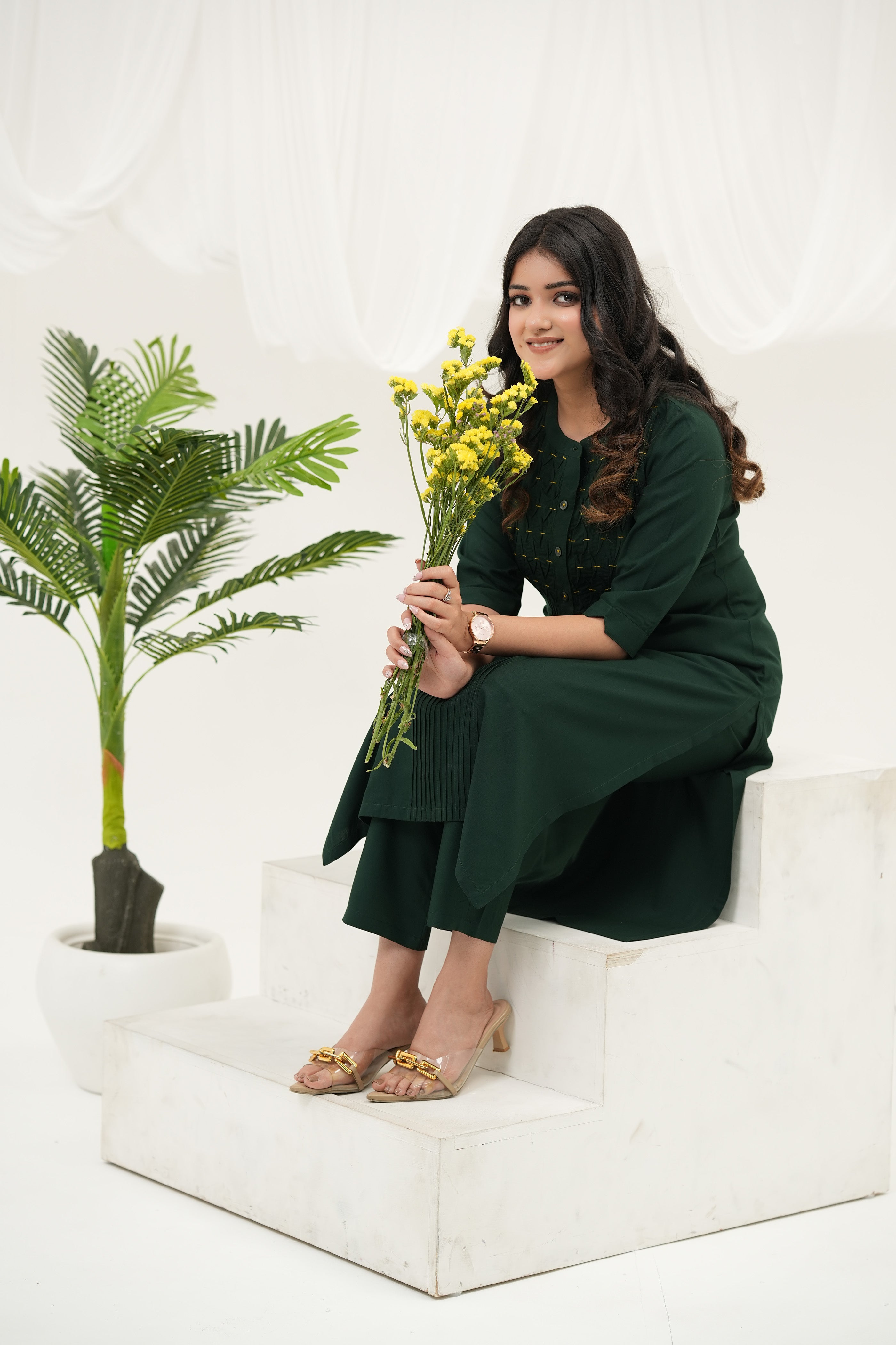 A model sitting at white stairs wearing a Emerald green kurta set for women with handcrafted beadwork, woven pintuck yoke, and 3/4 sleeves in breathable rayon viscose.