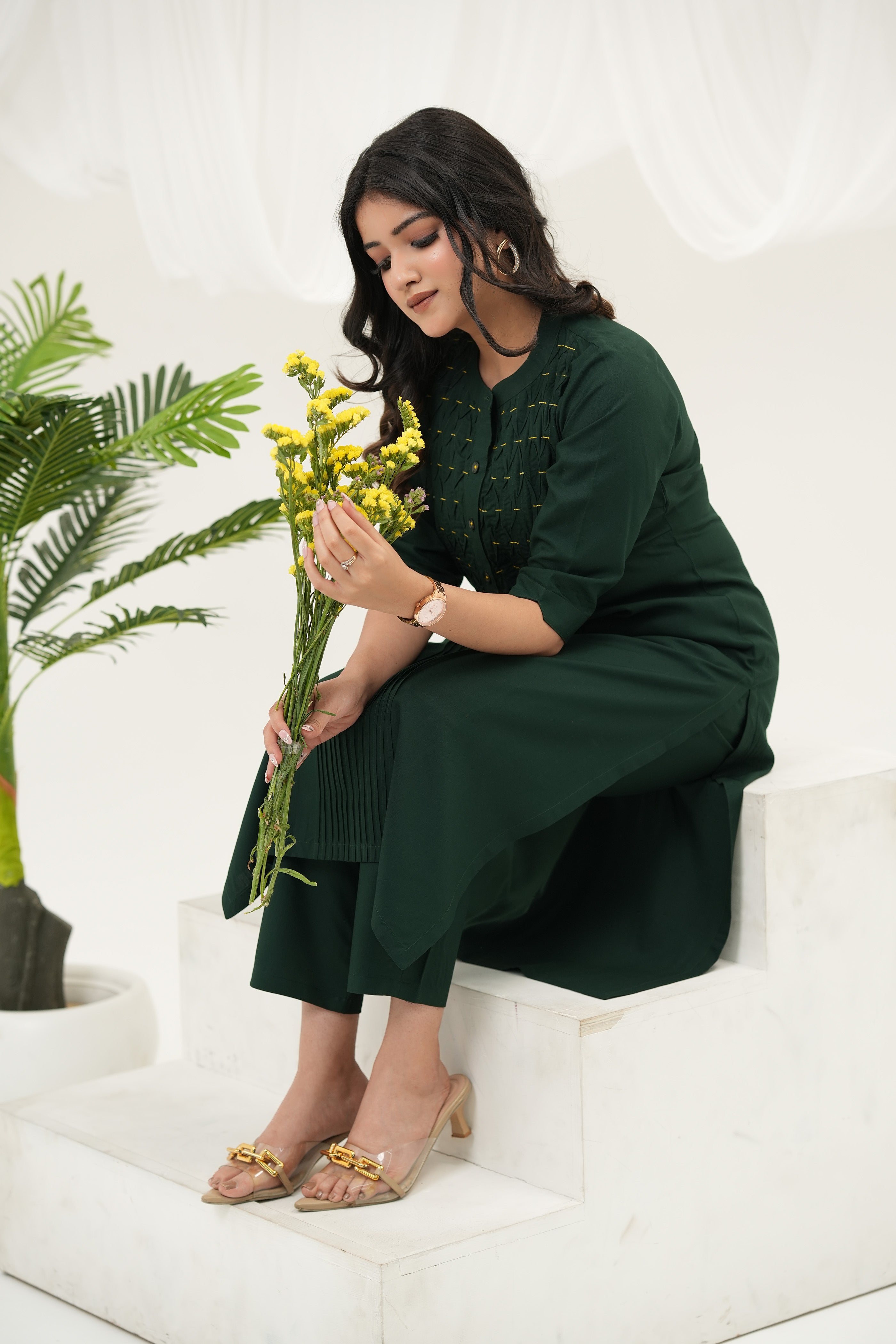 A model sitting at white stairs wearing a Emerald green kurta set for women with handcrafted beadwork, woven pintuck yoke, and 3/4 sleeves in breathable rayon viscose holding flowers in hands - Simroz.