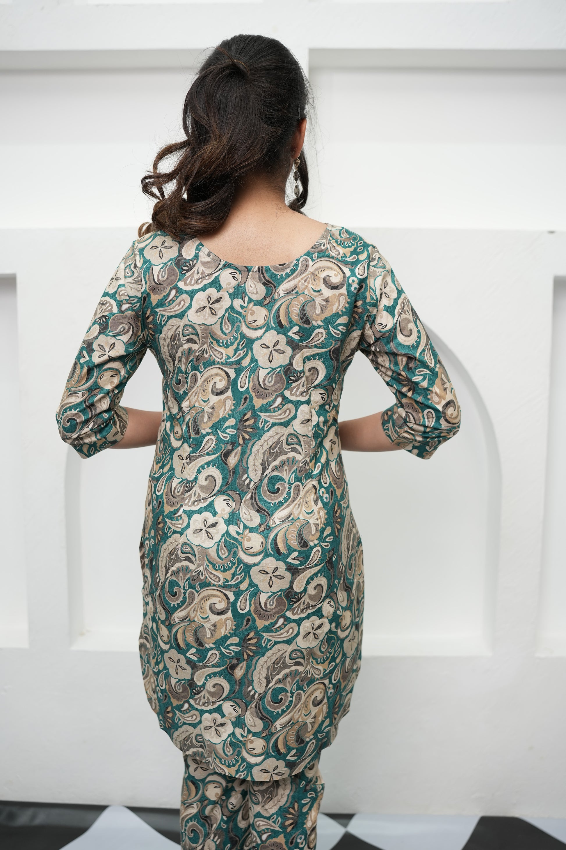 A woman wearing a paisley print tunic in deep teal, warm beige, and soft cream colors with three-quarter sleeves and a round neckline, back view . Simroz