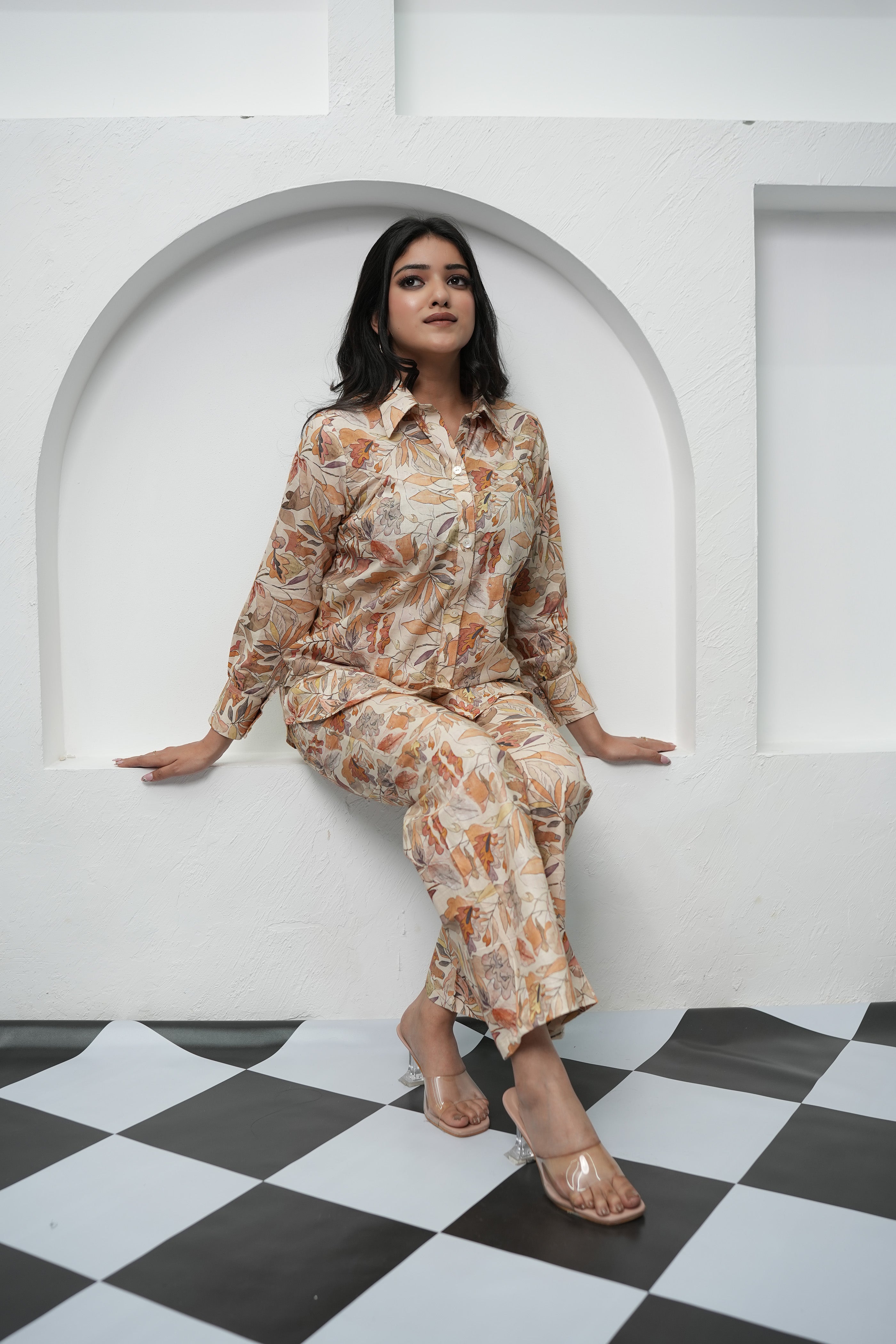 A woman sitting relaxed, wearing a beige and brown leaf patterned co-ord set consisting of a full sleeve top and pants - Simroz.