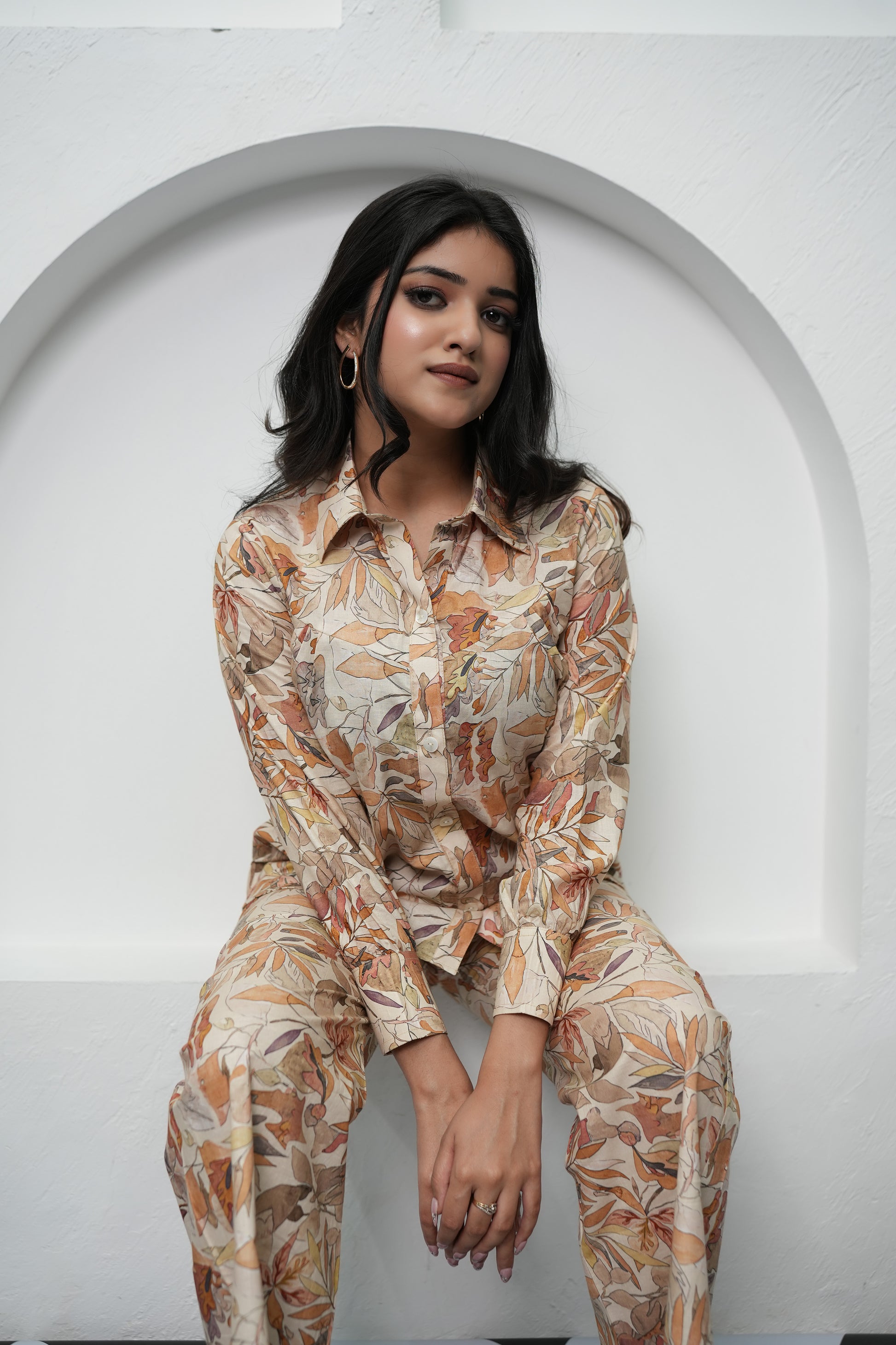 A woman sitting, wearing a beige and brown leaf patterned co-ord set consisting of a full sleeve top and pants - Simroz.